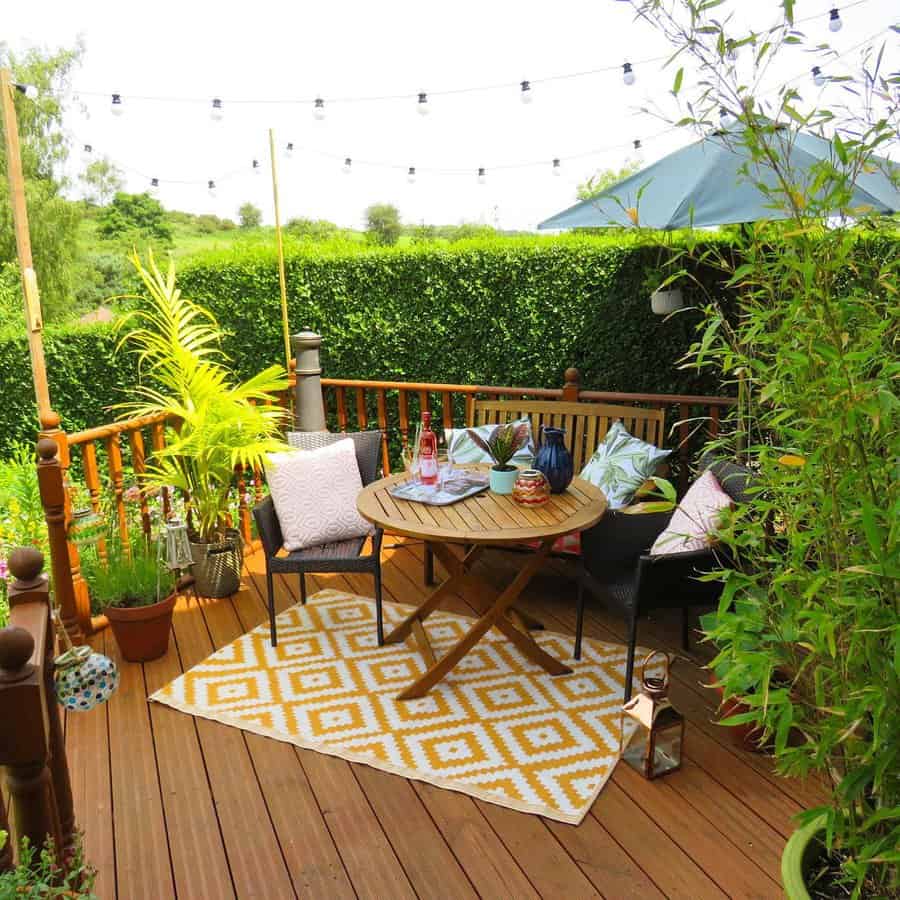 A cozy outdoor deck with wooden furniture, colorful cushions, a yellow patterned rug, string lights, and lush greenery around