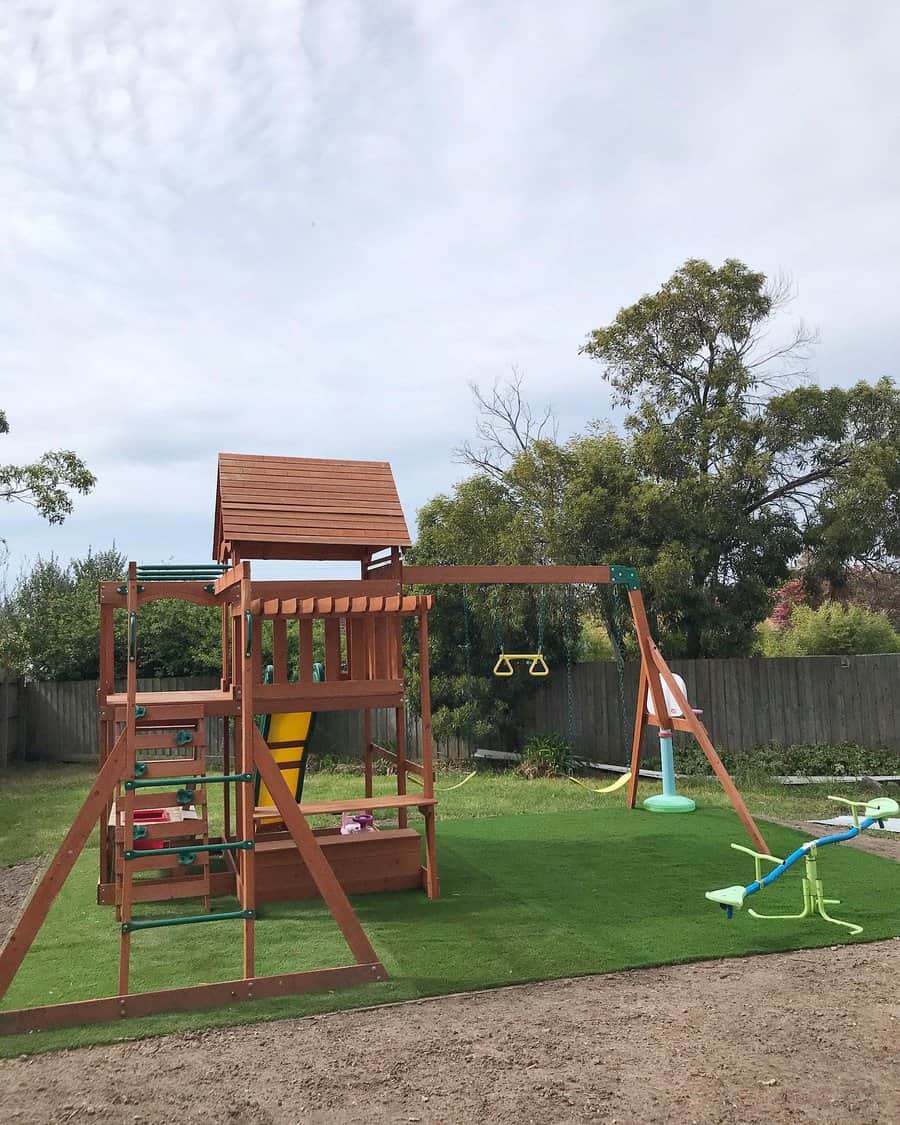 Children's playground set with slides and swings on artificial grass in a backyard, surrounded by trees and a wooden fence