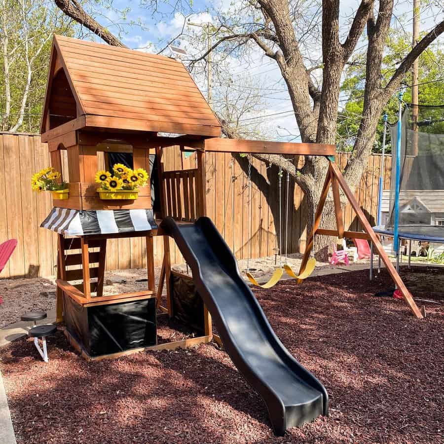 Wooden playset with slide, swings, and a small house featuring flower boxes set in a backyard with mulch ground and a trampoline nearby