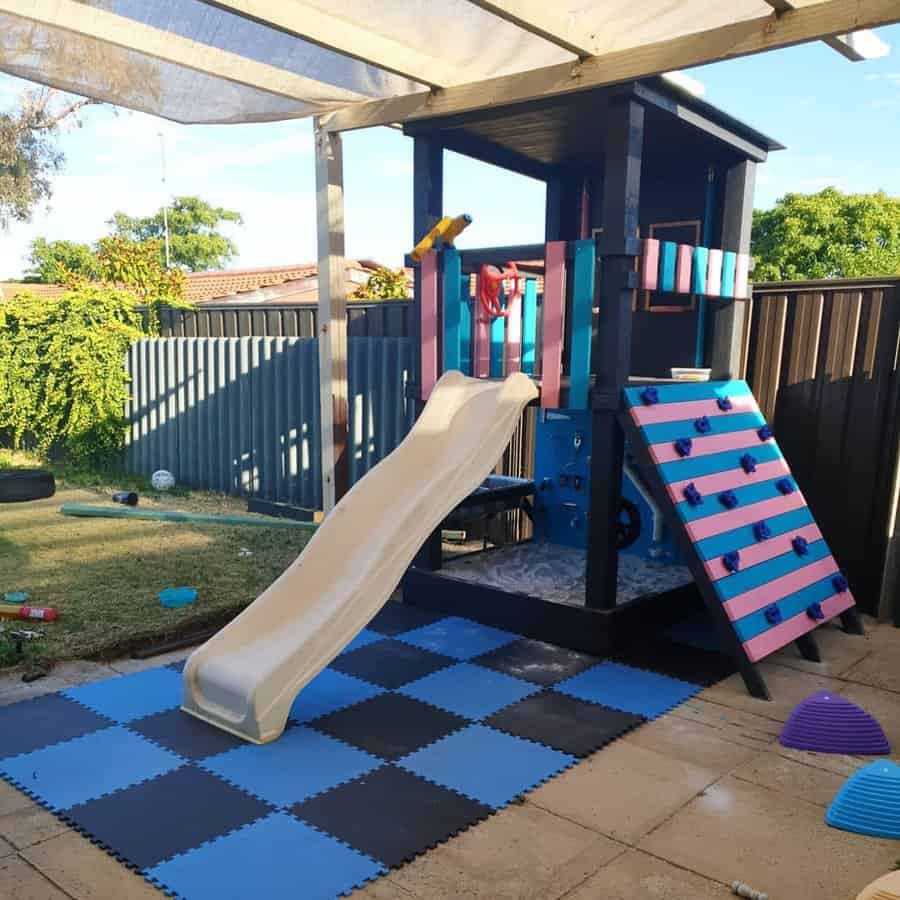 Children's play set with a beige slide, climbing wall, and sandbox, on a blue mat; it's outdoors with a fence and greenery nearby