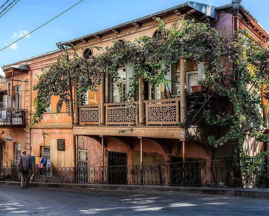 Vintage building with ornate balcony, vines, and weathered facade; man walking nearby on a sunny day