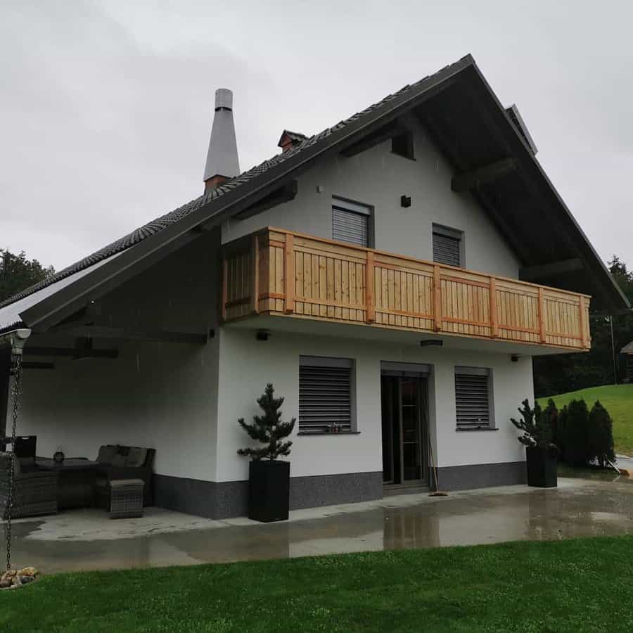 A modern white house with a wooden balcony and sloped roof, set in a rainy weather, featuring a small patio with furniture