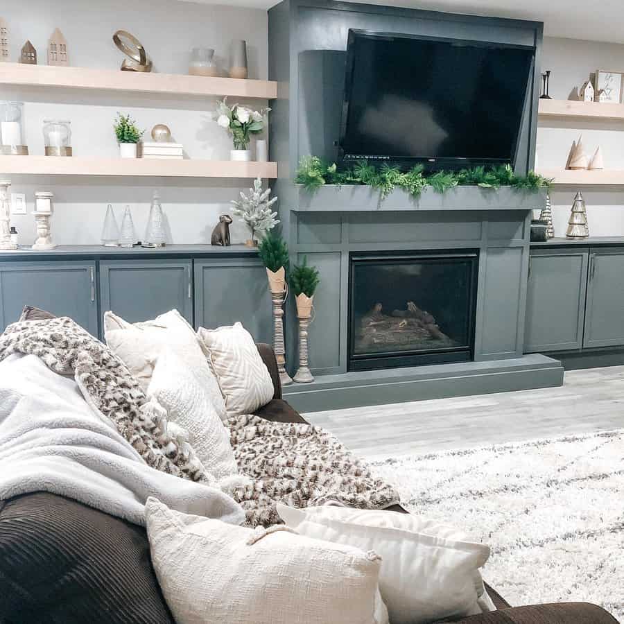 A cozy, modern living room featuring a dark green wooden fireplace mantel with a TV above, decorated with a greenery garland, and surrounded by stylish shelves and a comfy sofa