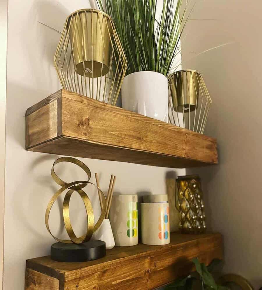 Wooden shelves with gold candle holders, potted grass, decorative jars, and abstract gold sculpture against a light wall