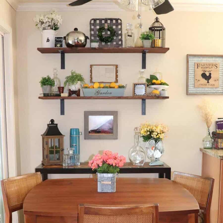 A cozy dining area with a wooden table, pink flowers, and three shelves featuring plants, decor, and framed art on the wall