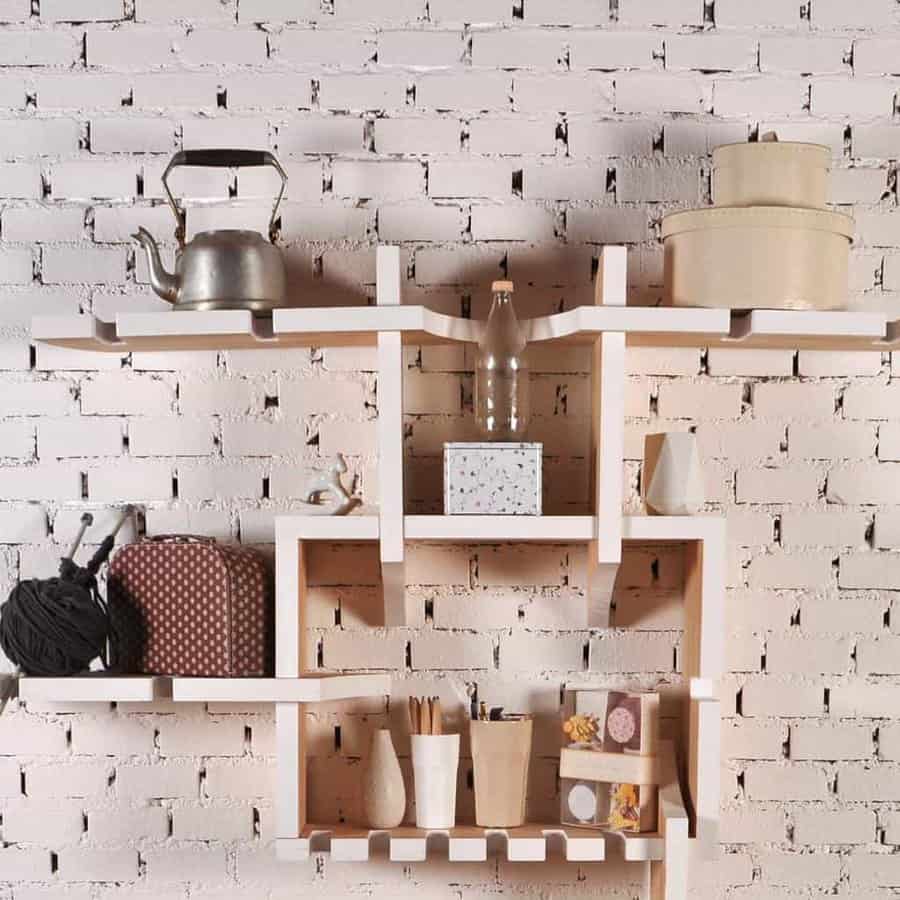 Modern white geometric shelves on a white brick wall, holding a kettle, boxes, yarn, and small decorative items