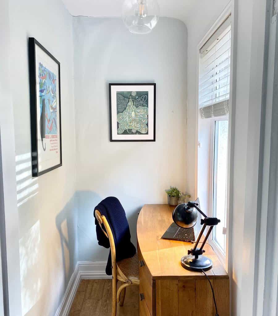 Cozy corner office with a wooden desk, lamp, laptop, and framed artwork. A chair with a blue blanket sits against a window with blinds