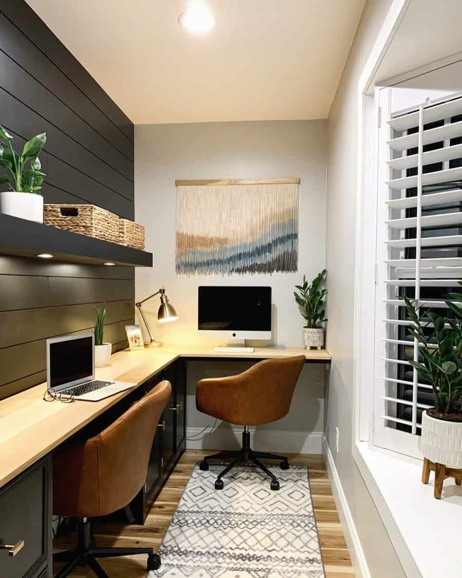 A cozy home office with two brown chairs, a computer on a wooden desk, plants, wall tapestry, and a large window with shutters