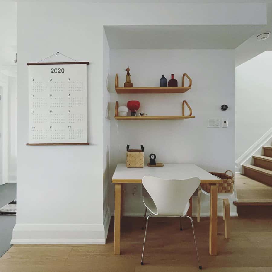 Minimalist workspace with a white desk, chair, wooden shelves, and a 2020 calendar on the wall, situated near a staircase