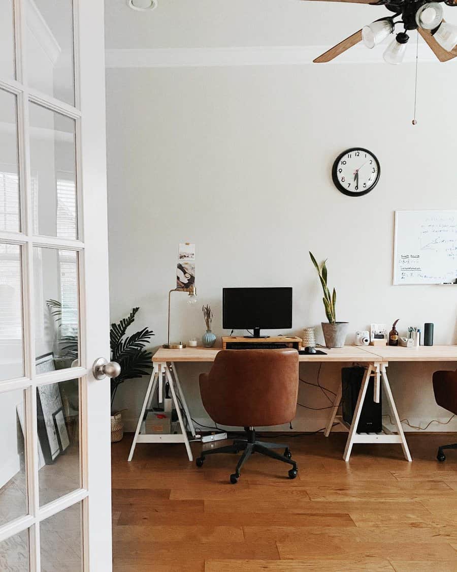 A home office with a brown chair, desk, computer monitor, plants, wall clock, and whiteboard; light wood floor and glass-paneled door