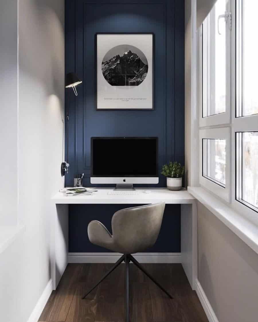 Minimalist workspace with a computer, gray chair, and a black lamp on a white desk against a blue wall, large windows on the right