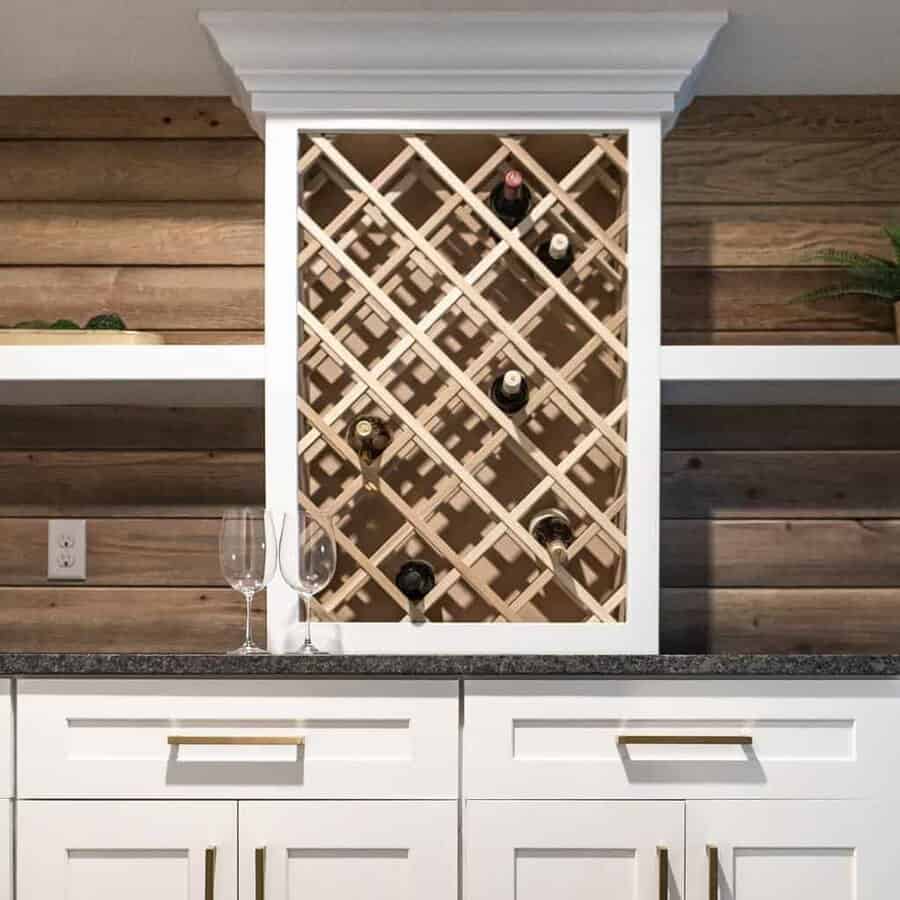 Wine rack with bottles on a wooden wall, two empty glasses on a countertop with white cabinets below