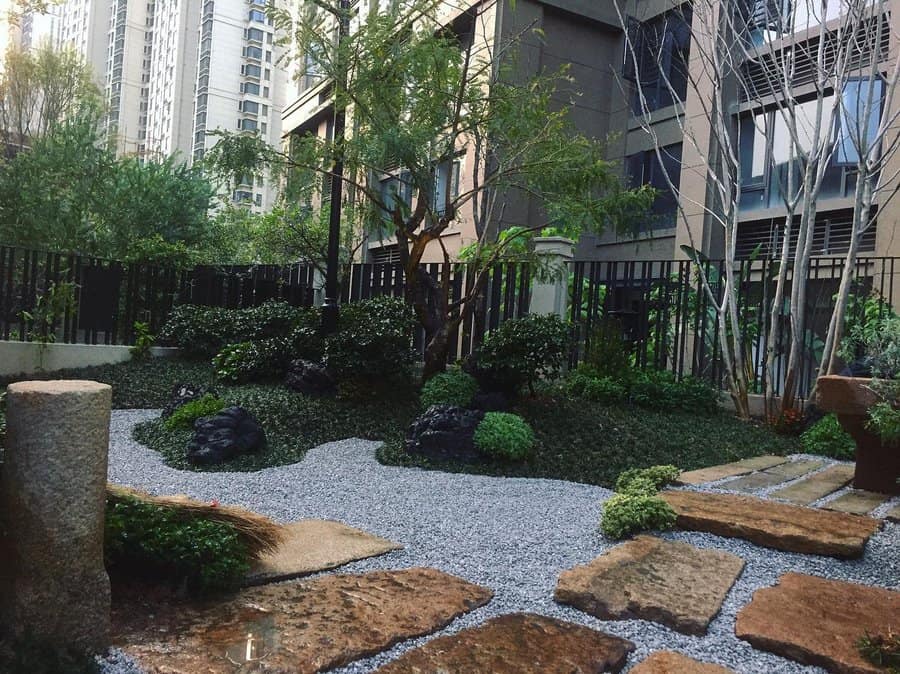 A tranquil Japanese-style garden with stone path, gravel, lush greenery, and background of modern buildings