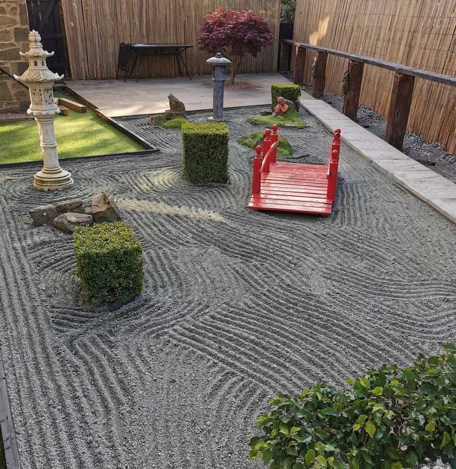 A small Japanese zen garden with raked gravel, a red wooden bridge, green shrubs, stone lantern, and decorative stones