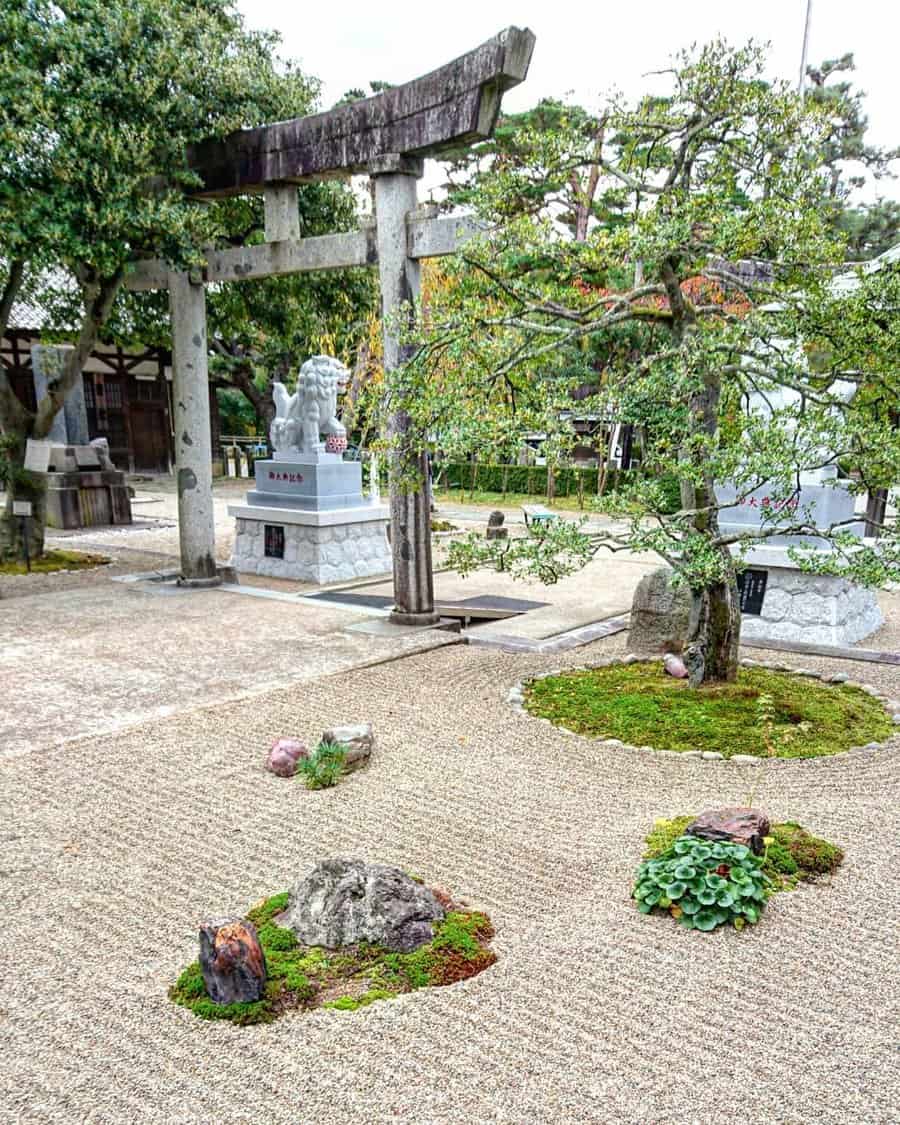 Japanese garden with a stone torii gate, a sculpted tree, raked gravel, moss islands, and a stone statue in the background