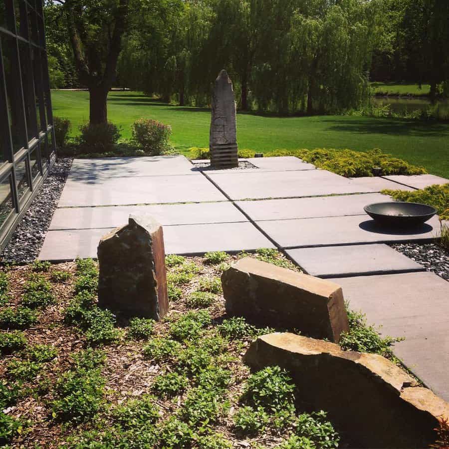 Modern garden with stone pathway, tall sculpture, lush greenery, and large windows reflecting trees