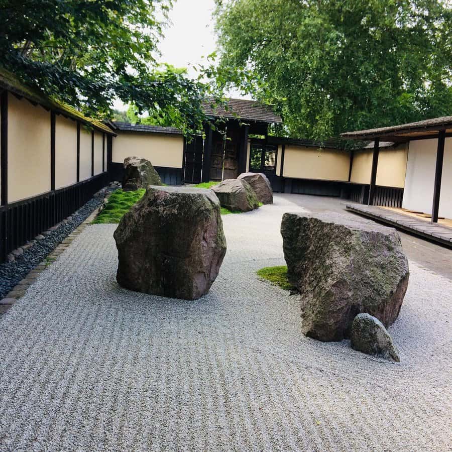 Japanese Zen garden with large rocks on raked gravel, surrounded by wooden walls and lush green trees; tranquil and minimalist