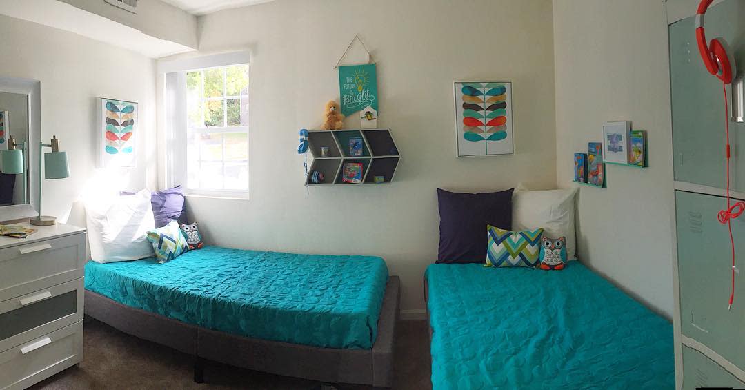 A cozy bedroom with two blue-covered beds, colorful decorations, wall art, a small shelf, and a window letting in natural light