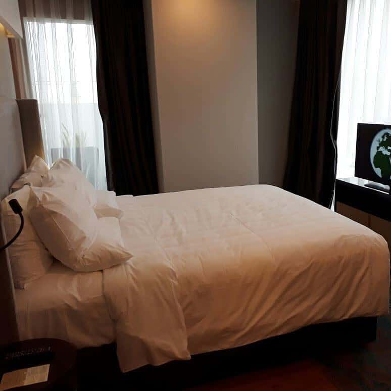 A neatly made bed in a dimly lit room with dark curtains and a TV showing an image of Earth on a dresser