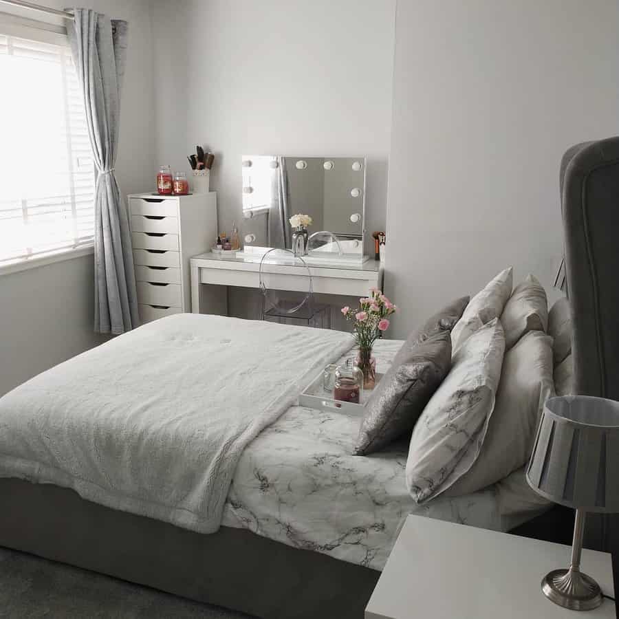 A small bedroom with a bed, white bedding, a makeup table with bulbs, gray curtains, and a small lamp on a bedside table