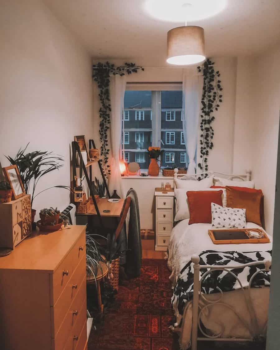 A small bedroom with a single bed, desk, and dresser; window with plant accents, warm lighting, and decorative pillows