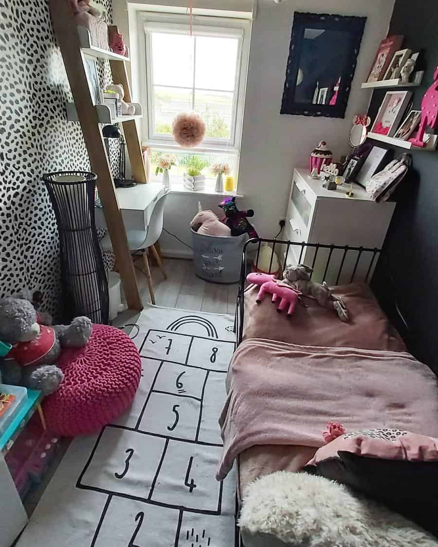 Small children's bedroom with a bed, hopscotch rug, desk by the window, stuffed animals, and wall shelves featuring pink and leopard print accents