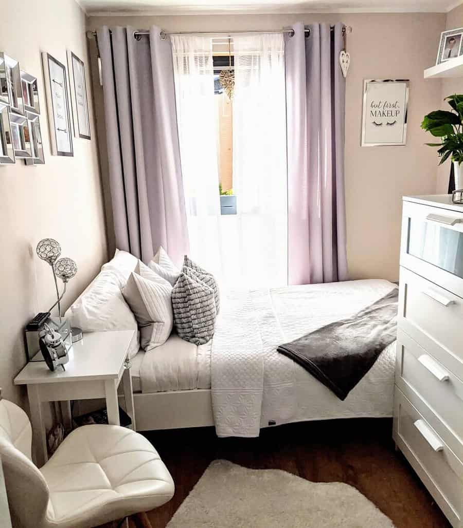 Cozy small bedroom with white bedding, gray curtains, pillows, and a small desk with a chair; wall art and a plant decorate the room