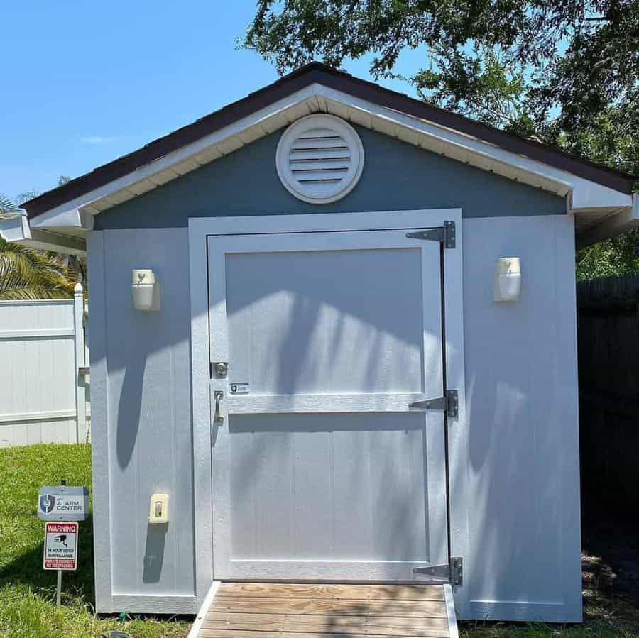 A small gray shed with a white door and roof, vent above, and ramp leading up, surrounded by grass, trees, and a white fence