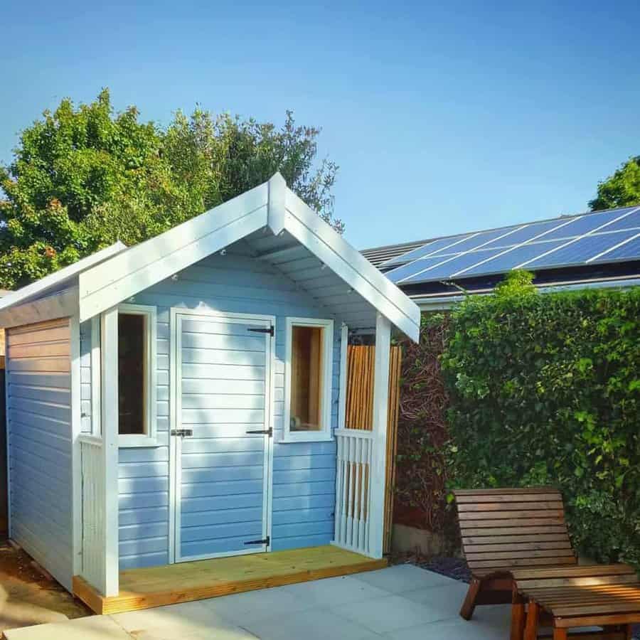 A small blue wooden shed with a peaked roof is next to a building with solar panels, surrounded by greenery and a wooden chair