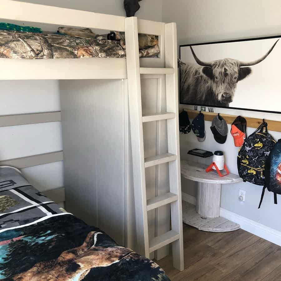 Cozy bedroom with a bunk bed, camouflage bedding, a ladder, a cow portrait, hats and backpacks on hooks, and a small table