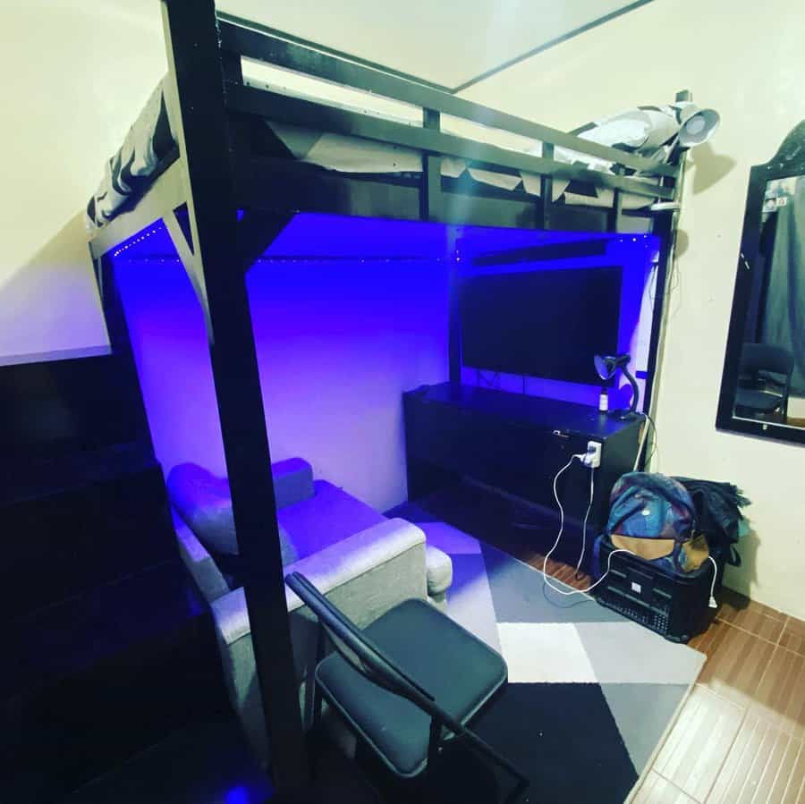 Loft bed with a blue LED-lit workspace underneath, featuring a desk, TV, chair, small couch, and shelves in a compact room