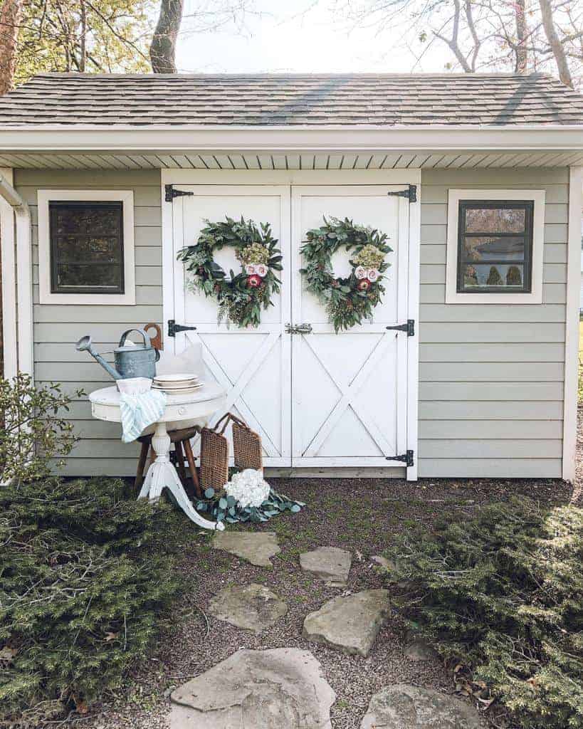 Small gray shed with wreaths on white doors, stone path, and vintage garden decor with flowers and watering cans in the foreground