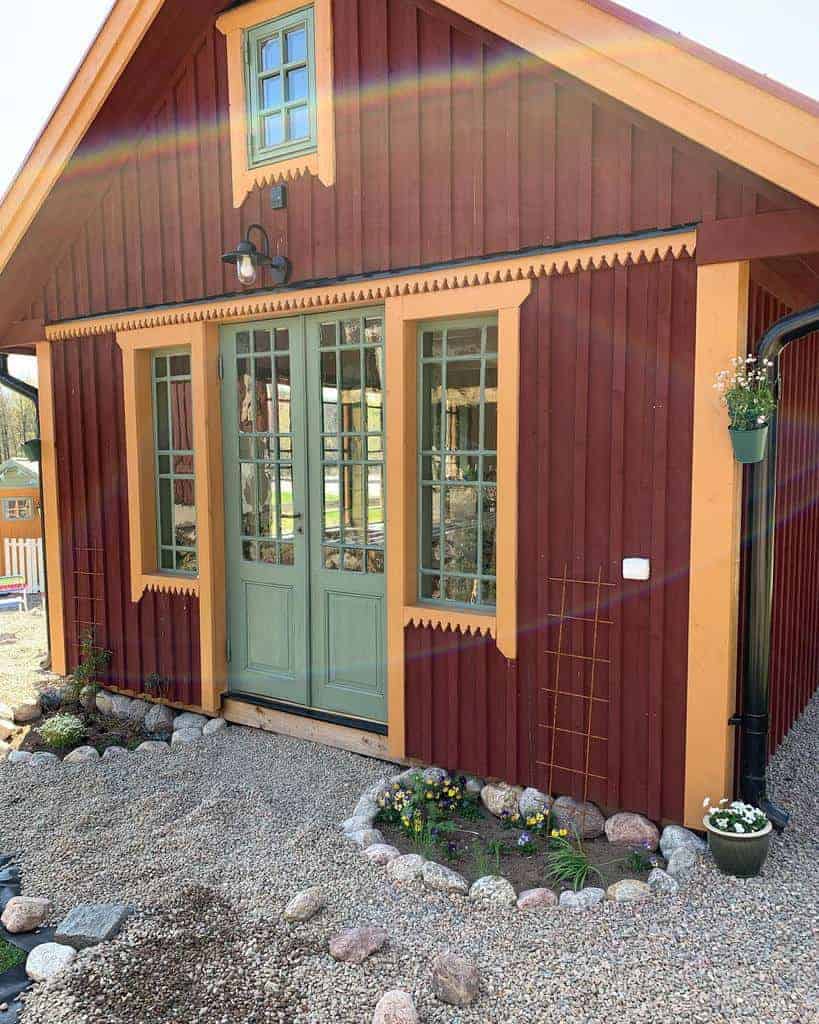 A small red and orange wooden cottage with decorative trim, a green door, and plants and stones in the garden bed in front