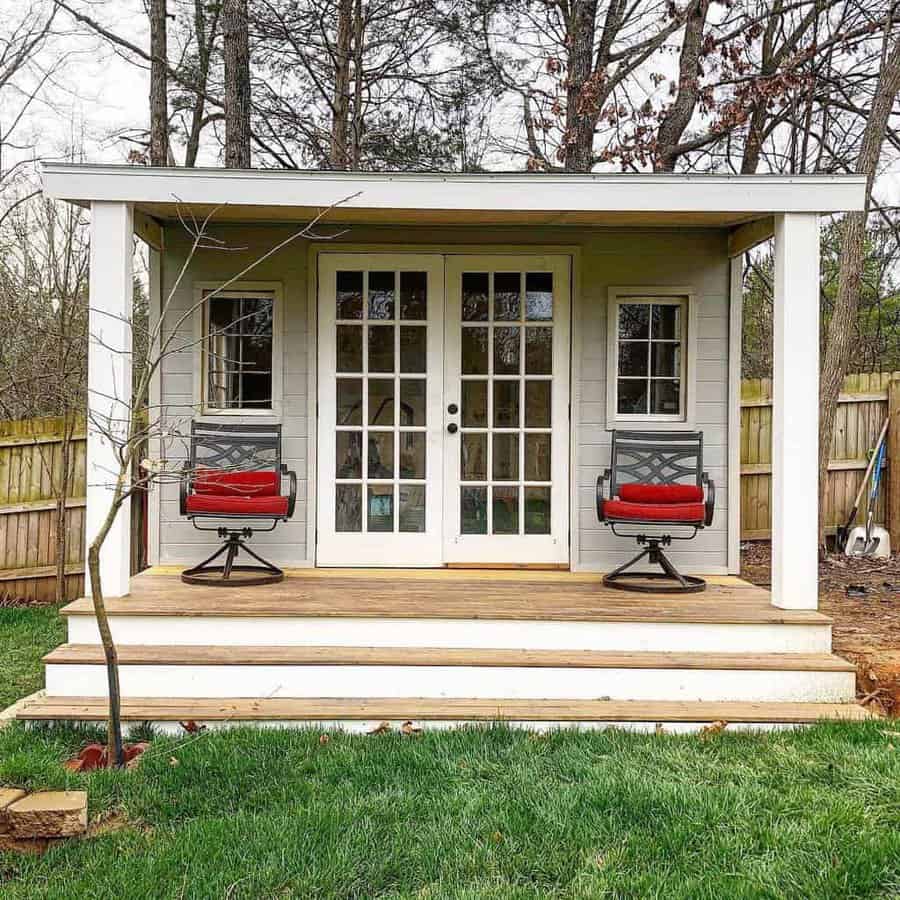 A small gray shed with a porch, two red-cushioned chairs, and a glass door, set in a grassy yard with a wooden fence and trees