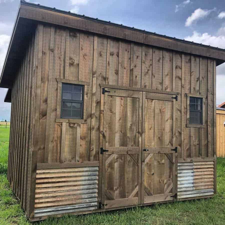 Wooden shed with metal accent panels, double doors, and two small windows, standing in a grassy area under a partly cloudy sky