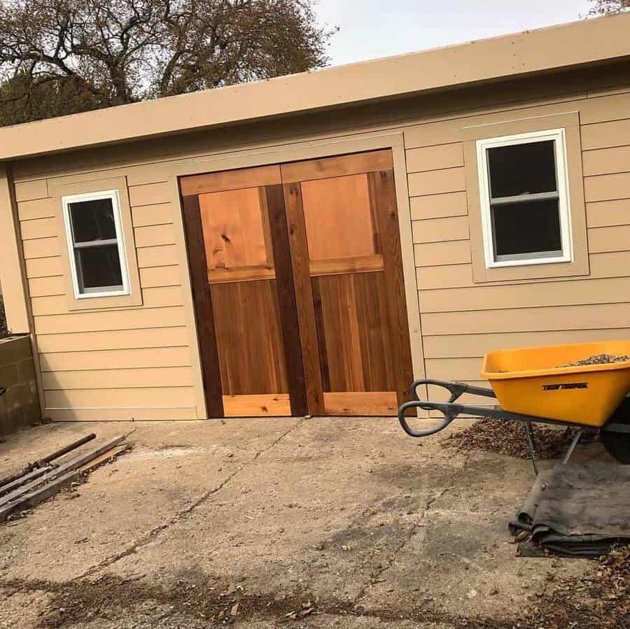 A beige shed with wooden double doors, two windows, and a yellow wheelbarrow in front on a concrete surface