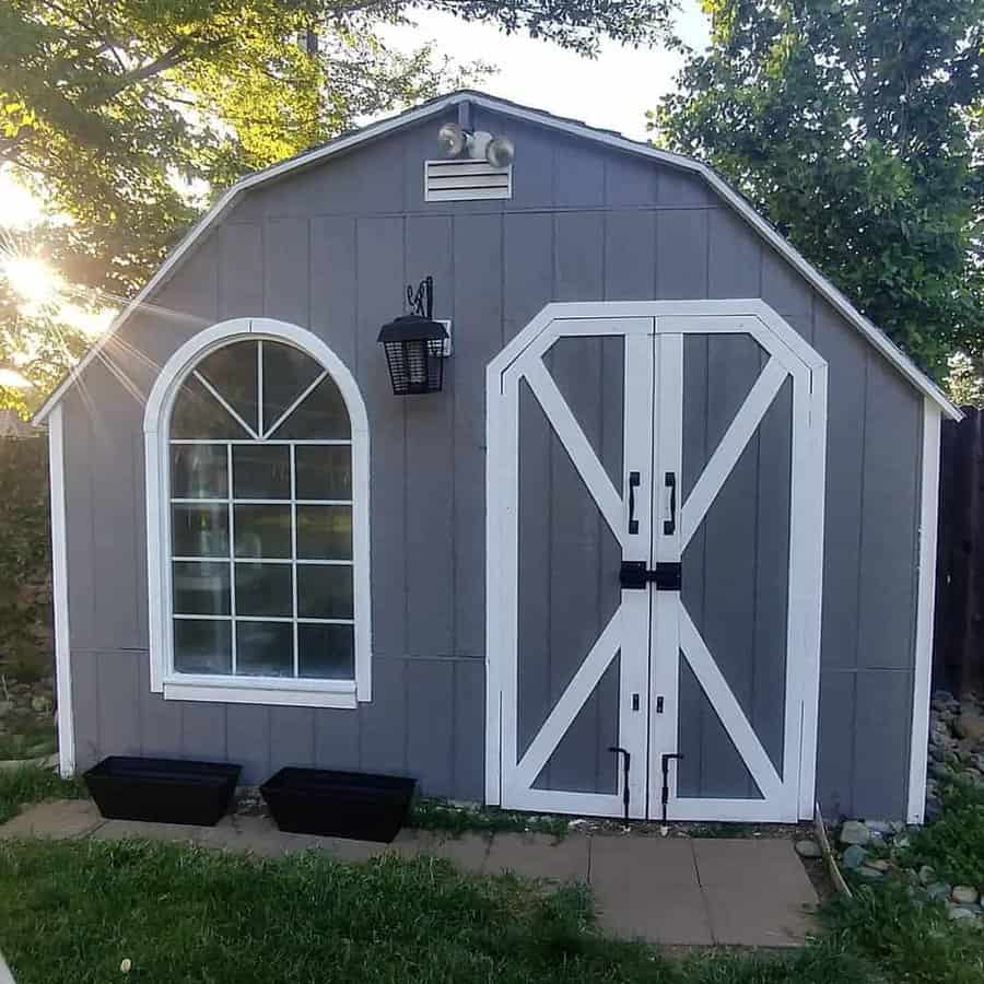 A gray shed with a barn-style door, adjacent arched window, and exterior light, surrounded by grass and trees