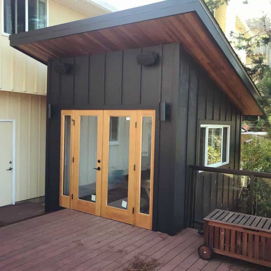 Modern garden shed with angled wooden roof, large glass doors, and dark exterior, set on a wooden deck with outdoor bench