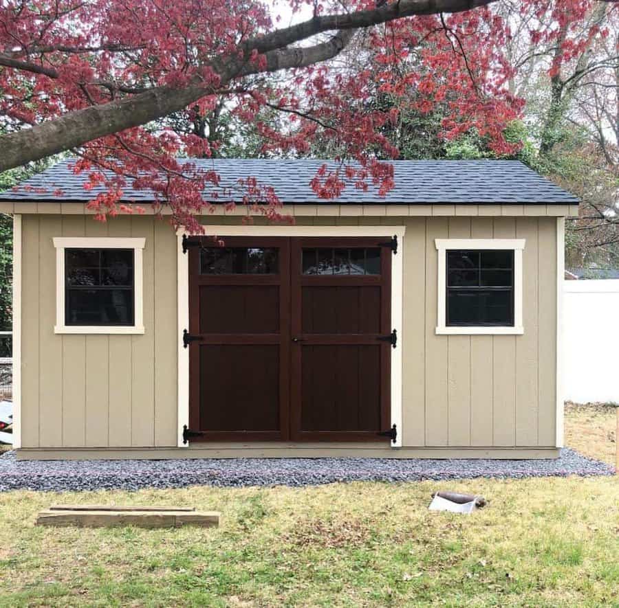 A beige garden shed with a dark brown double door and two windows under a tree with red leaves