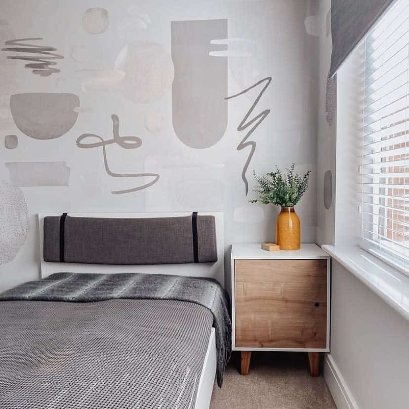 A minimalist bedroom featuring abstract wall art, a gray bed, wooden nightstand with a potted plant, and light filtering through window blinds