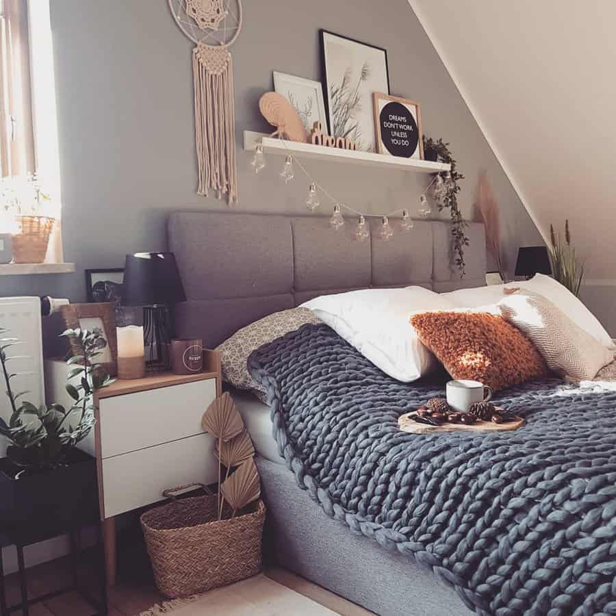 Cozy bedroom with a gray bed, chunky knit blanket, orange pillow, plants, and a dreamcatcher; soft lighting and decor accents