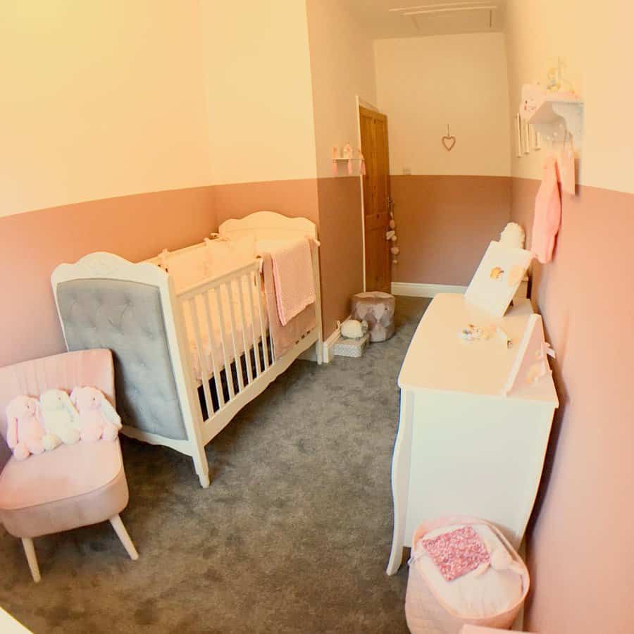 Cozy nursery with pink and white decor, featuring a crib, plush chair, dresser, and wall shelves with toys; carpeted floor