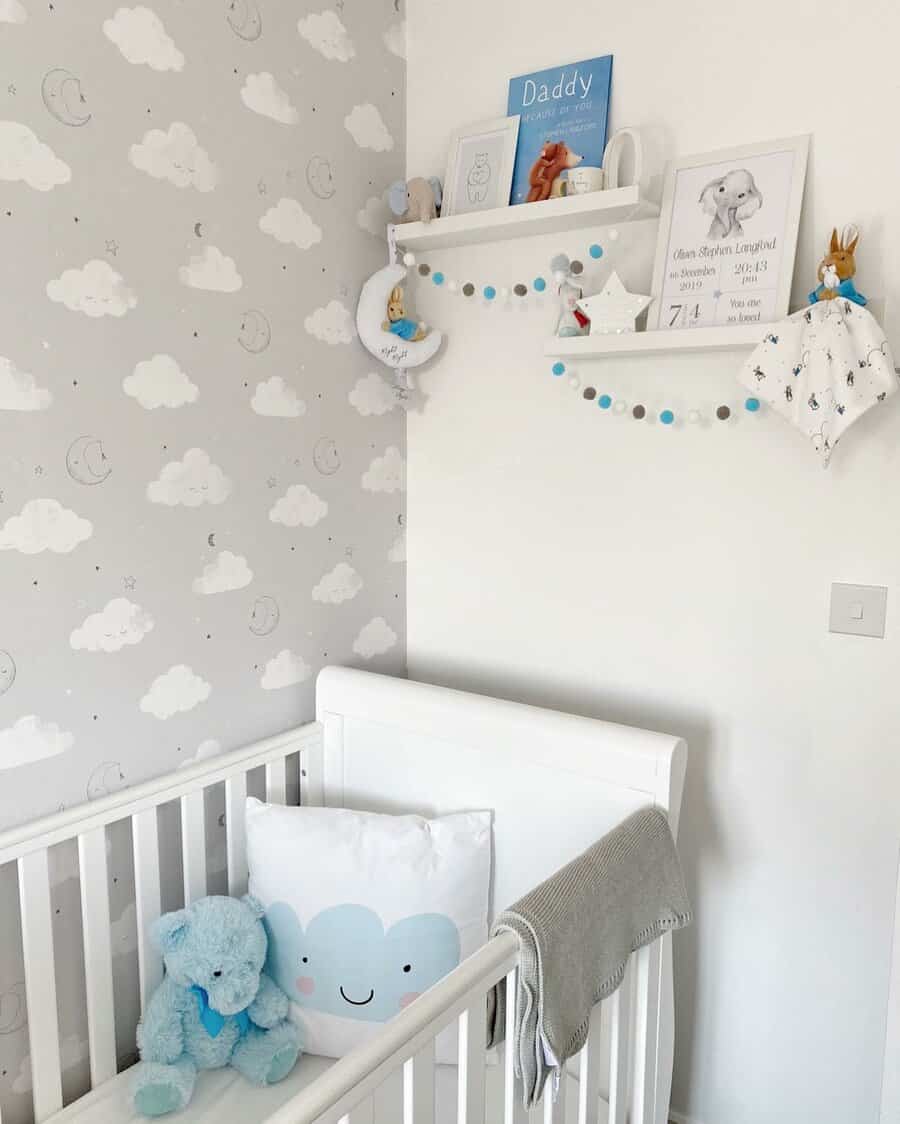 A white baby crib with a cloud pillow and blue teddy bear, cloud-patterned wallpaper, shelves with books, toys, and wall decor above