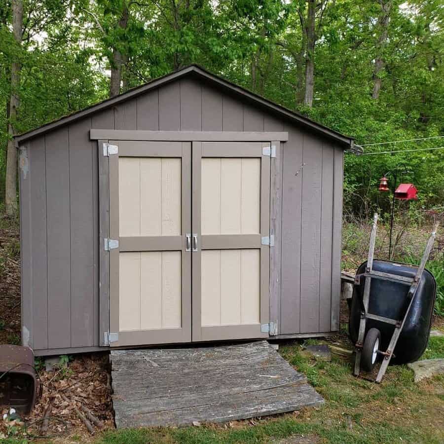 Rustic gray shed with beige double doors, silver metal hinges, and handles, set in a wooded backyard with a weathered wooden ramp