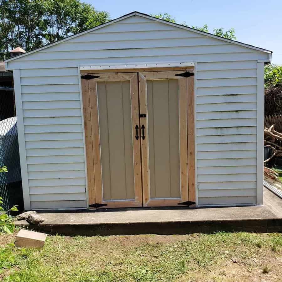 White shed with double doors and black hinges, set on a concrete slab in a grassy yard, with trees in the background