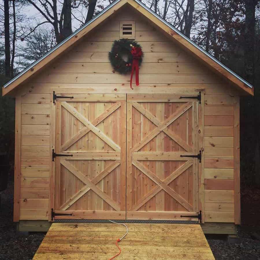 Wooden shed with double doors, decorated with a holiday wreath above, sits amidst trees; an orange extension cord lies on the ramp