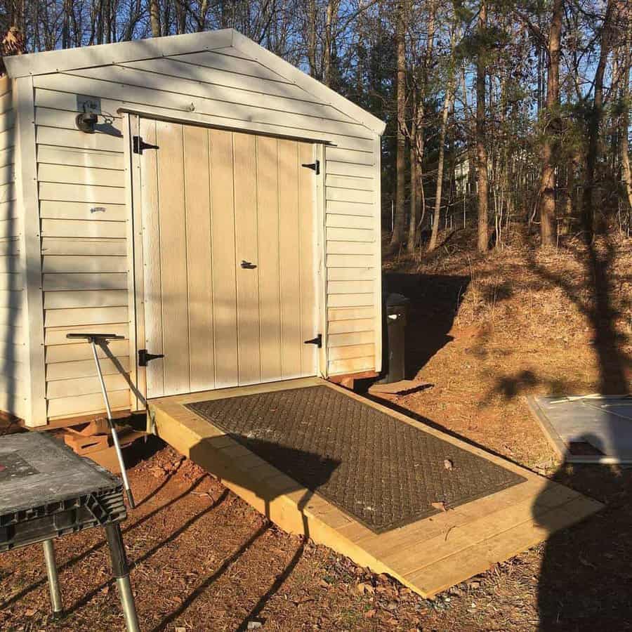 Beige shed with a double door, black hinges, and a sturdy wooden ramp with a non-slip mat, set in a wooded area with sunlight