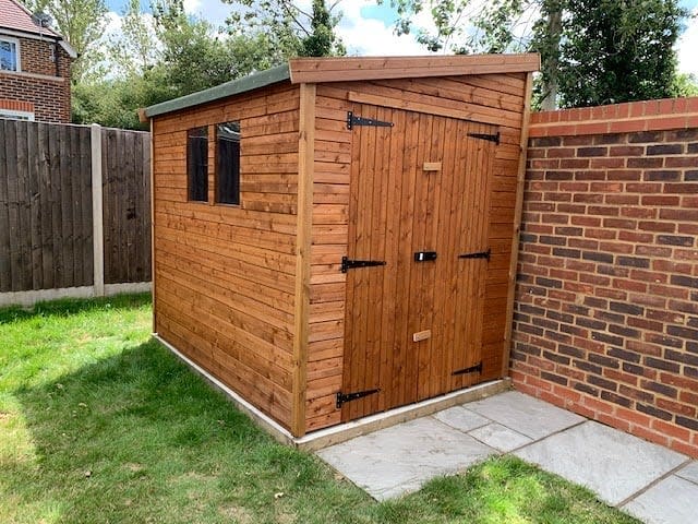 A wooden shed with a small window and a double door, situated in a grassy backyard near a brick wall and fence