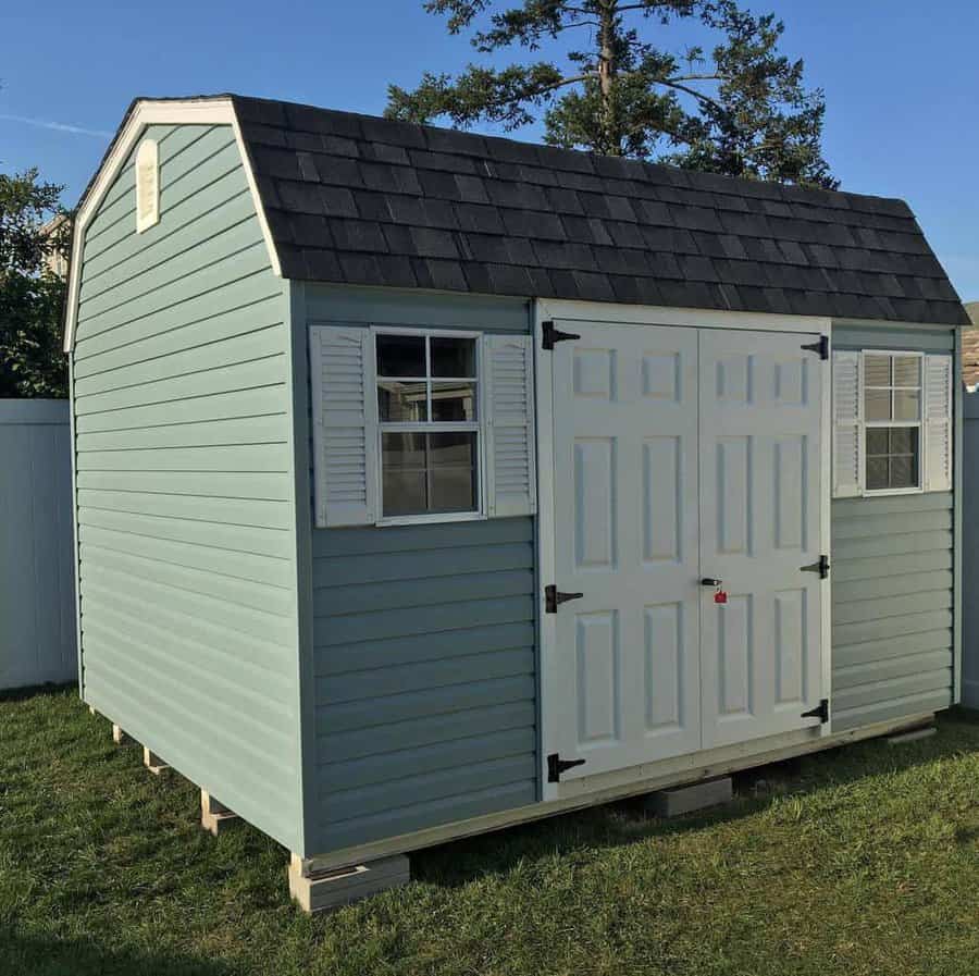 A small, light blue shed with a shingled roof, white double doors, and window shutters, set on a grassy lawn