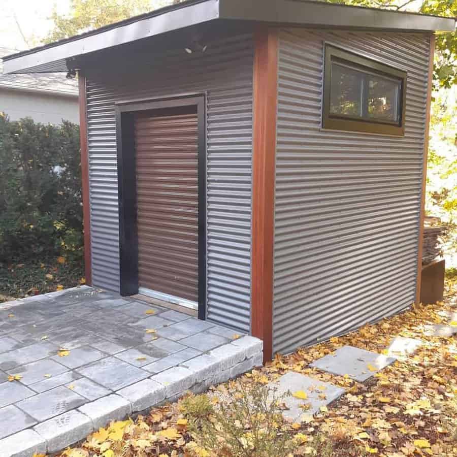 Modern gray corrugated metal shed with a brown door, surrounded by autumn leaves on a paved patio area